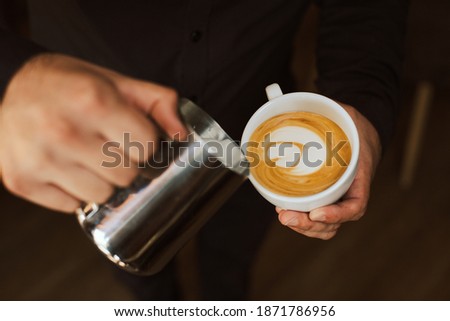 Closeup image of male barista hands pouring steamed milk in to a ceramic coffee cup, creating a fresh latte art cappuccino.