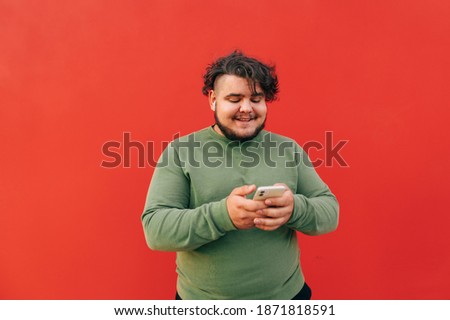Smiling cheerful young bearded guy is enjoying the music on his headphones, typing the messages on his smartphone and feeling excited using the device.
