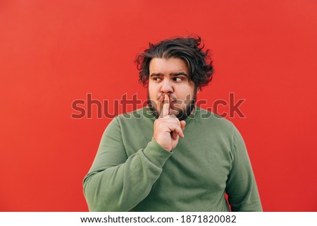 Funny attractive young bearded hispanic man is showing a shh gesture by touching his mouth with his finger, looking aside, expressing silence sign, standing on a red background.