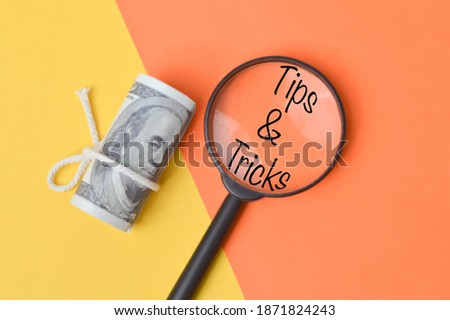 Top view of money banknote and magnifying glass written with text TIPS and TRICKS over yellow and orange background. Business concept.