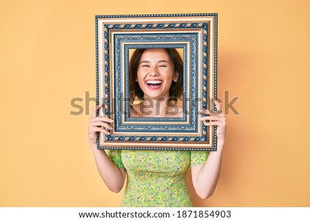 Young beautiful woman holding empty frame smiling and laughing hard out loud because funny crazy joke. 