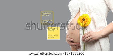 A pregnant woman in a white shirt holds a yellow gerbera flower on a gray isolated background. Colors of the 2021 year yellow and grey