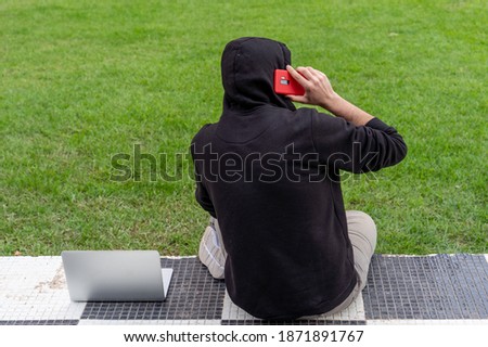 A young man wearing a hoodie sitting in a park with his laptop talking on the phone