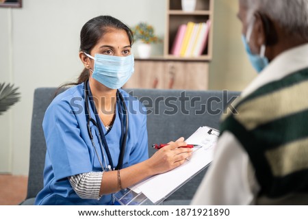Shoulder shot, young doctor or nurse writing prescription during home visiting to sick senior man while both worn face mask due to coronavirus covid-19 pandemic as safety measures. Royalty-Free Stock Photo #1871921890