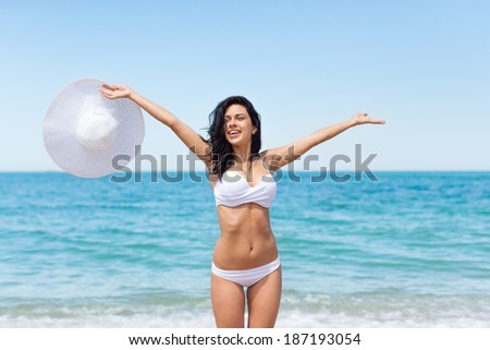 woman happy excited smile on beach ocean hold raised arms hands up, white hat and bikini swimsuit, young girl summer vacation holiday on sea sunny day blue sky
