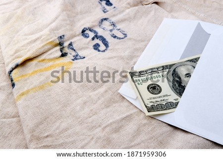 One hundred dollar bill lies in a white mail envelope and partially peeps out of it. Corrupt bribe in paper packaging. The table is covered with a dense light fabric with a number printed on top.