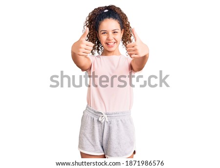 Beautiful kid girl with curly hair wearing casual clothes approving doing positive gesture with hand, thumbs up smiling and happy for success. winner gesture. 