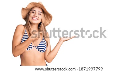 Young beautiful blonde woman wearing bikini and hat amazed and smiling to the camera while presenting with hand and pointing with finger. 