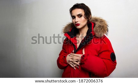 Stylish young girl smile in the winter street in old town wearing red down jacket, black gloves and grey knitted hat. Winter holiday concept Girl in a red down jacket with red lipstick and wet hair.
