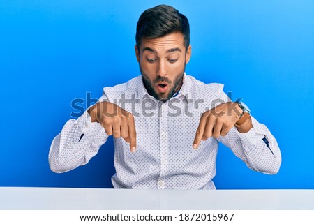 Handsome hispanic man wearing business clothes sitting on the table pointing down with fingers showing advertisement, surprised face and open mouth 