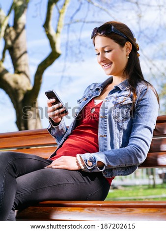 Photo of a beautiful young woman using a smartphone while sitting in a park on a bench.