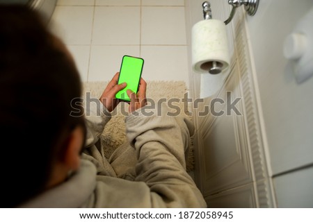 Man at Bathroom Sitting on Toilet Using Phone With Green Mock-up. a person will not melt with the phone. Tapping on Center Screen. green screen. dependence.        