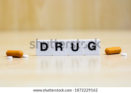A row of small white plastic tiles, containing the letters forming the word drug, placed on a table together with tablets and capsules, to represents the concept of drug use or topic.