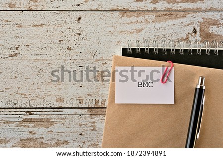 There is a piece of paper with "BMC" written on it. It was an abbreviation for Business Model Canvas. It was on top of white damaged wood table with a envelope and a notebook.