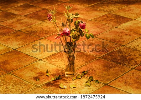 Beautiful close up photo of dried pink roses in autumn bouquet standing in transparent glass vase on rustic old tiles floor. Dried leaves fallen from the flowers. Soft light, blurry background