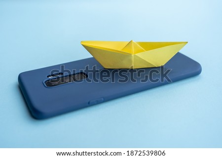 A closeup shot of a blue case smartphone with a yellow paper boat  on a blue surface