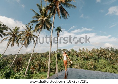 Woman walking on edge of infinity pool in white  bikini, hold in hands  straw hat in jungle around trees. Vacation concept