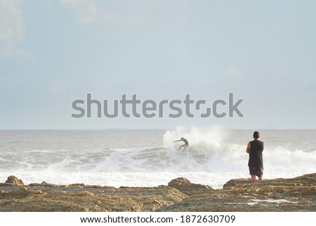 Man standing on a rocky coastline watching a surfer ride a big wave.