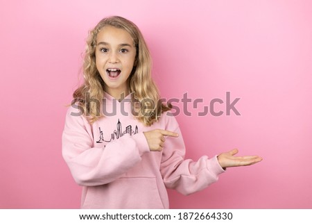 Young beautiful child girl standing over isolated pink background surprised, showing and pointing something that is on her hand