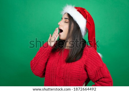 Young beautiful woman wearing Christmas Santa hat over isolated green background bored yawning tired covering mouth with hand. Restless and sleepiness.
