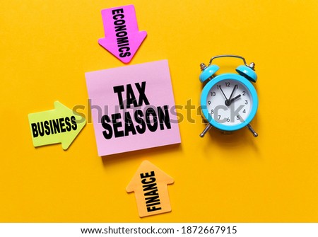 Finance and economics concept. On a yellow background, a blue alarm clock, multi-colored paper arrows with inscriptions and paper for notes on which the text is written - TAX SEASON