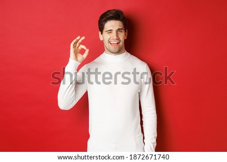 Portrait of charismatic beaded guy in white sweater, assure you, winking and showing okay sign to guarantee good quality, praise excellent choice, standing over red background