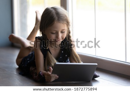 Close up little girl lying on warm floor near window, using tablet, enjoying leisure time with gadget, playing game, browsing apps, interested child looking at device screen, watching video