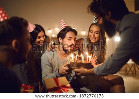 Make a wish Handsome young man in his twenties, wearing party cap, blowing burning candles on his birthday cake surrounded by happy, cheerful, smiling friends who came to his birthday party Royalty-Free Stock Photo #1872776173