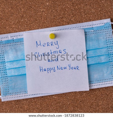 Merry Christmas and Happy New Year. White square sticker with writing in blue gel pen and pinned on medical mask and corkboard. Christmas wishes.