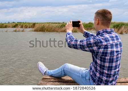 Man in blue shirt taking photography of landscape. He is sitting on wooden dock next to lake.