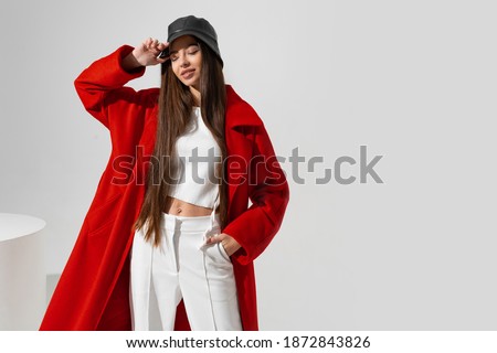 Fashion portrait of beautiful skinny   model in stylish  hat and red coat   posing on white background in studio.  Wearing crop top and  pants.