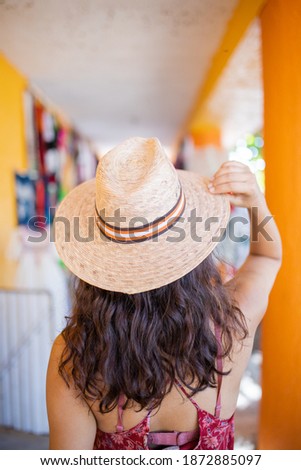 Brunette woman wearing red dress and straw hat standing next to hat rack. Young woman trying on different straw hats. Summer beauty and fashion Royalty-Free Stock Photo #1872885097