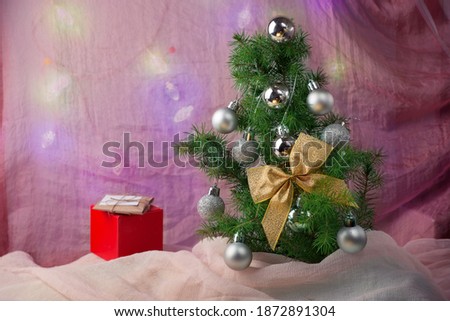 Small spruce tree with Christmas decorations and a gold bow on the pink background with a red present box