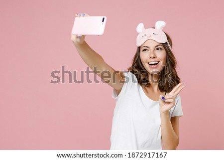 Funny young woman in white pajamas home wear sleep mask showing victory sign doing selfie shot on mobile phone resting at home isolated on pink background studio portrait. Relax good mood concept
