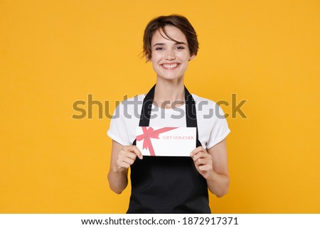 Smiling cheerful young female woman 20s barista bartender barman employee in white t-shirt apron posing holding in hand gift certificate isolated on yellow color wall background studio portrait