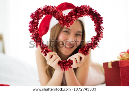 Selected focus of heart shape frame with blurred image of Asian Attractive  young Wolman on bed wearing red Santa hat and red gift box during Christmas holiday. new normal concept.