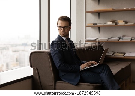 Serious young Caucasian businessman sit in chair in office work on laptop look in distance planning or thinking. Pensive male director or boss use computer at workplace. Business vision concept. Royalty-Free Stock Photo #1872945598