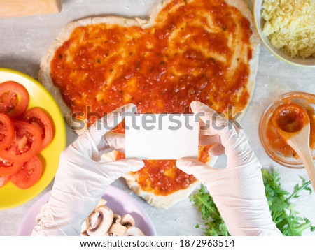 Hands holding empty business card on hear shape pizza dough with tomato sauce background. Ingredients for cooking pizza with dough in heart shape