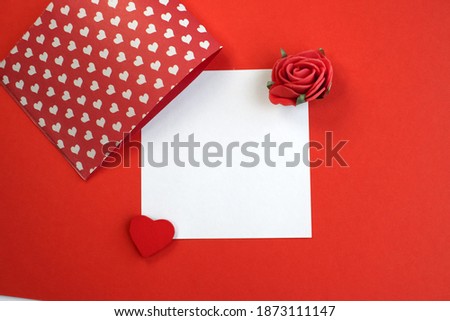 A white square leaf with a heart and a rose lies on a red background. Holiday background template with copy space