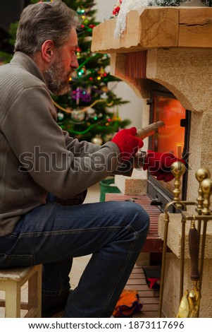 New Year by the fireplace. A man with a beard lays a log in the burning fireplace. In the background, New Year lights are lit on the tree.

