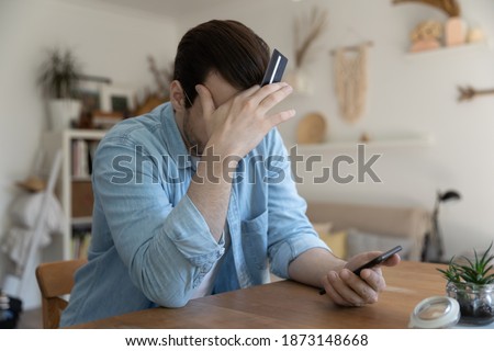 Close up depressed young man holding credit card and smartphone, covering face with hand, having problem with online banking service, cyber fraud, loss money, scam concept Royalty-Free Stock Photo #1873148668