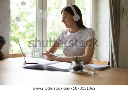 Smiling young woman wearing headphones using laptop, making video call, studying with teacher, learning language online, sitting at desk at home, female student watching webinar, listening lecture