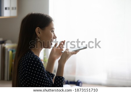 Close up side view young woman recording voice message, holding phone near mouth, sitting at desk at home, chatting online, making call, talking by speakerphone, activating assistant on smartphone