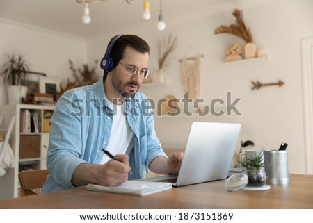 Focused man wearing headphones writing notes, looking at laptop screen, student watching webinar, listening to lecture, home office, businessman engaged in online negotiations, conference