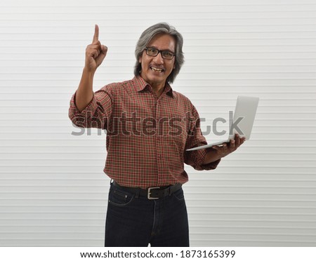Happy asian senior businessman in red casual shirt and blue jeans holding laptop computer and pointing finger up standing over white wall background, Business confident and success concept