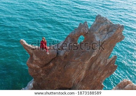 A beautiful girl with long dark hair and red clothes is sitting astride a huge rock in the shape of a dragon on the background of the sea. The concept of active recreation.