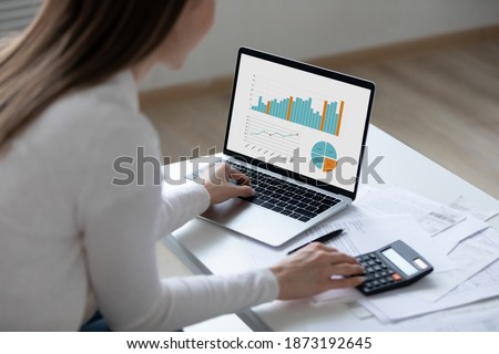 Close up rear view young woman using laptop and calculator, working on project, checking financial documents, sitting at table at home, looking at computer screen with diagrams, calculating bills Royalty-Free Stock Photo #1873192645