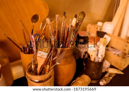 Different types of brushes for oil and acrylic painting, palette knives stand upright in clay vases in the artist's studio.