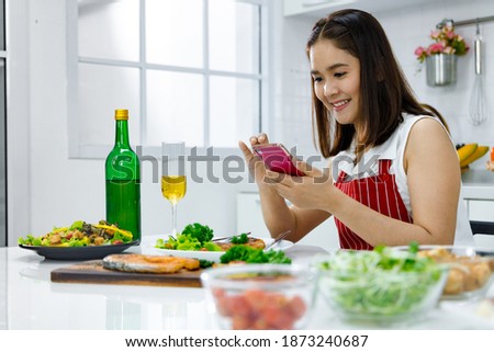 Asian woman in red apron taking a picture of food she make with mobile phone. Concept woman preparing meals at home.