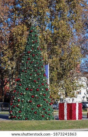 Artificial Christmas tree with bright red and gold balls and a decorative gift in an open area on a sunny December day
